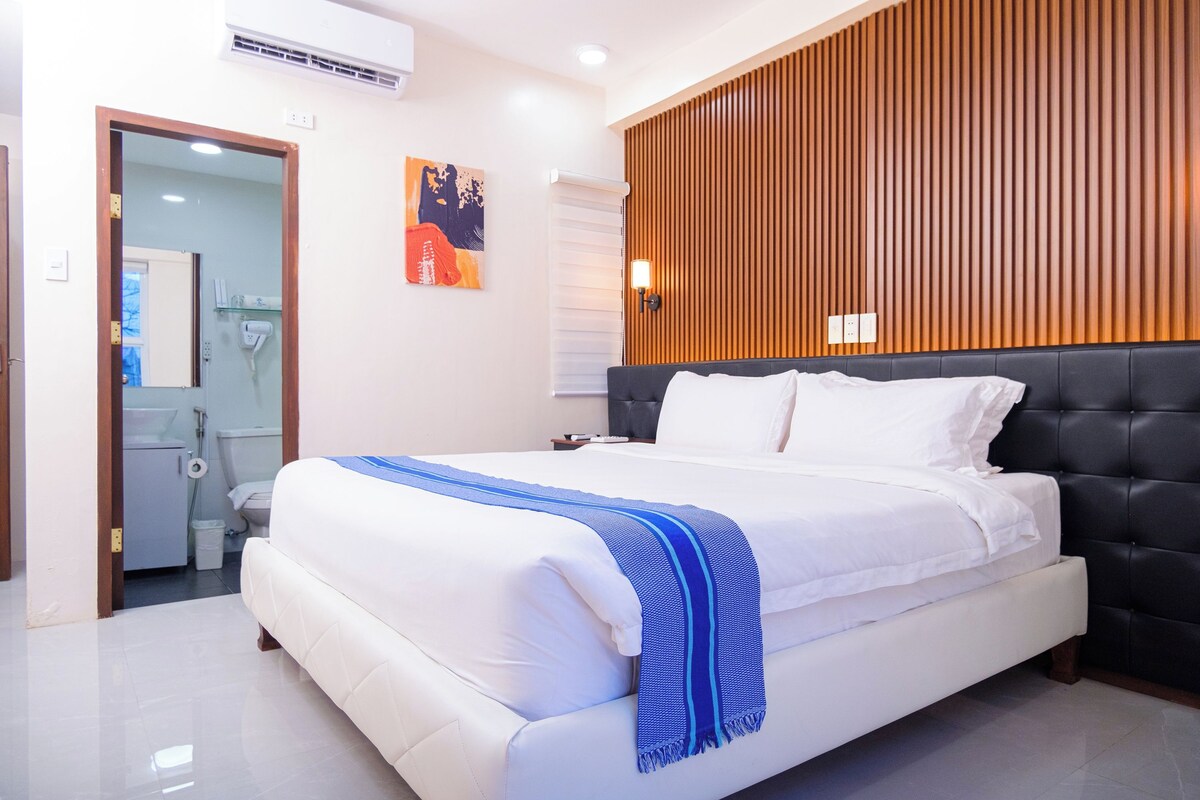Ein modernes Hotelzimmer mit einem großen Bett, einer Wandpaneele aus Holz und einem durch eine geöffnete Tür einsehbaren Badezimmer.