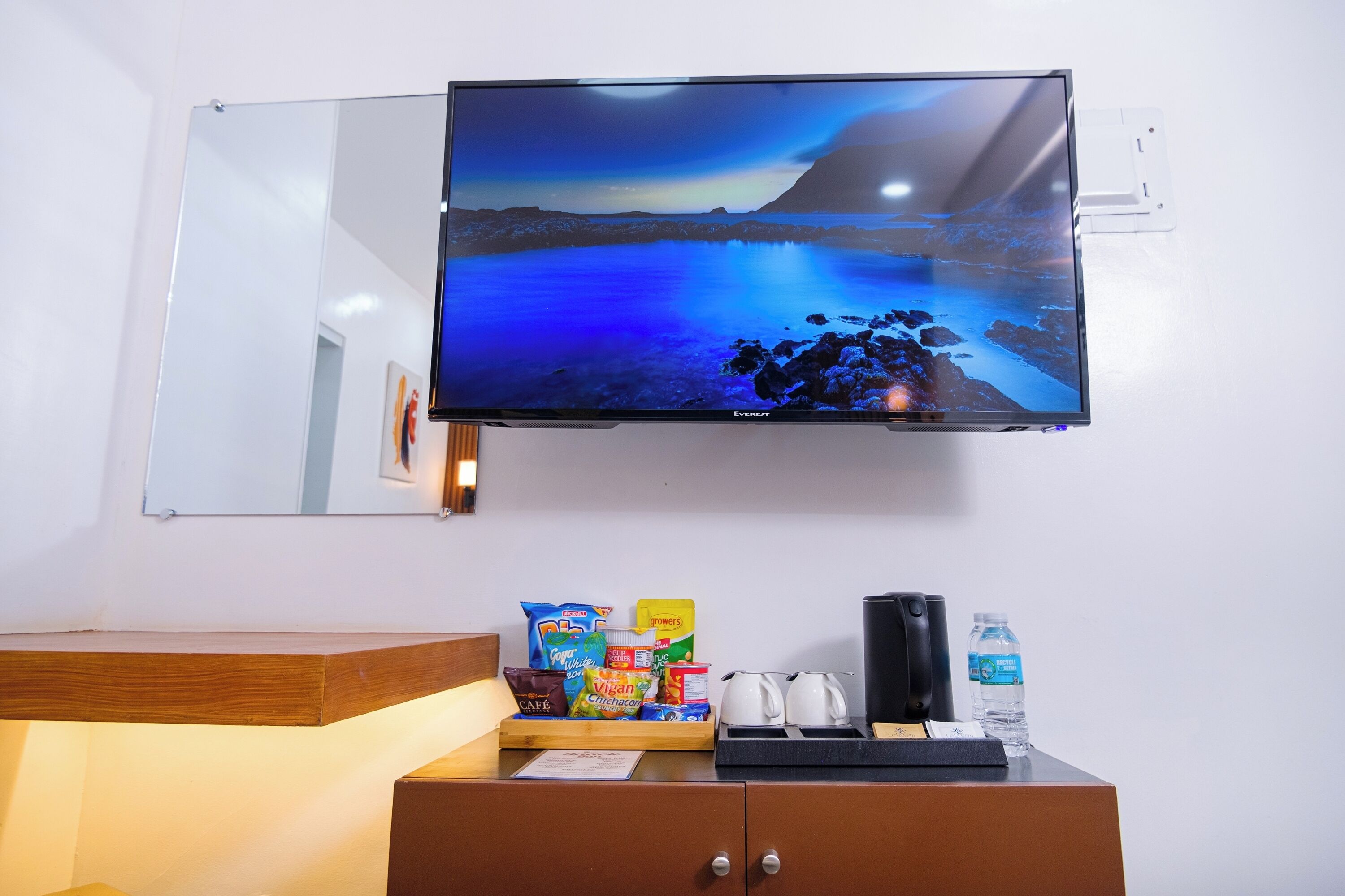 A flat-screen TV mounted on a wall displaying a night scene. Below, a wooden counter holds snacks, a coffee maker, and a water bottle.