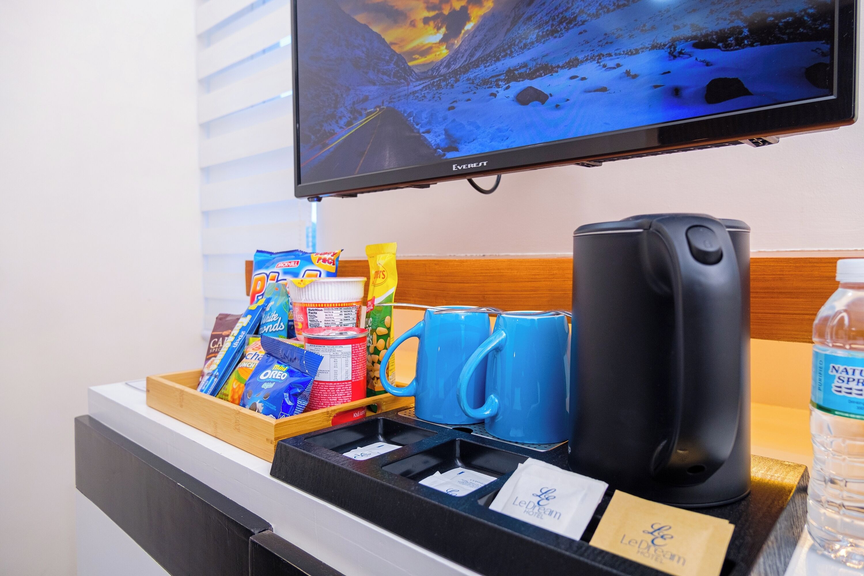 Zona de recepció d'hotel amb una televisió que mostra un paisatge nevós, una màquina de cafè, tasses blaves, aperitius i una ampolla d'aigua.