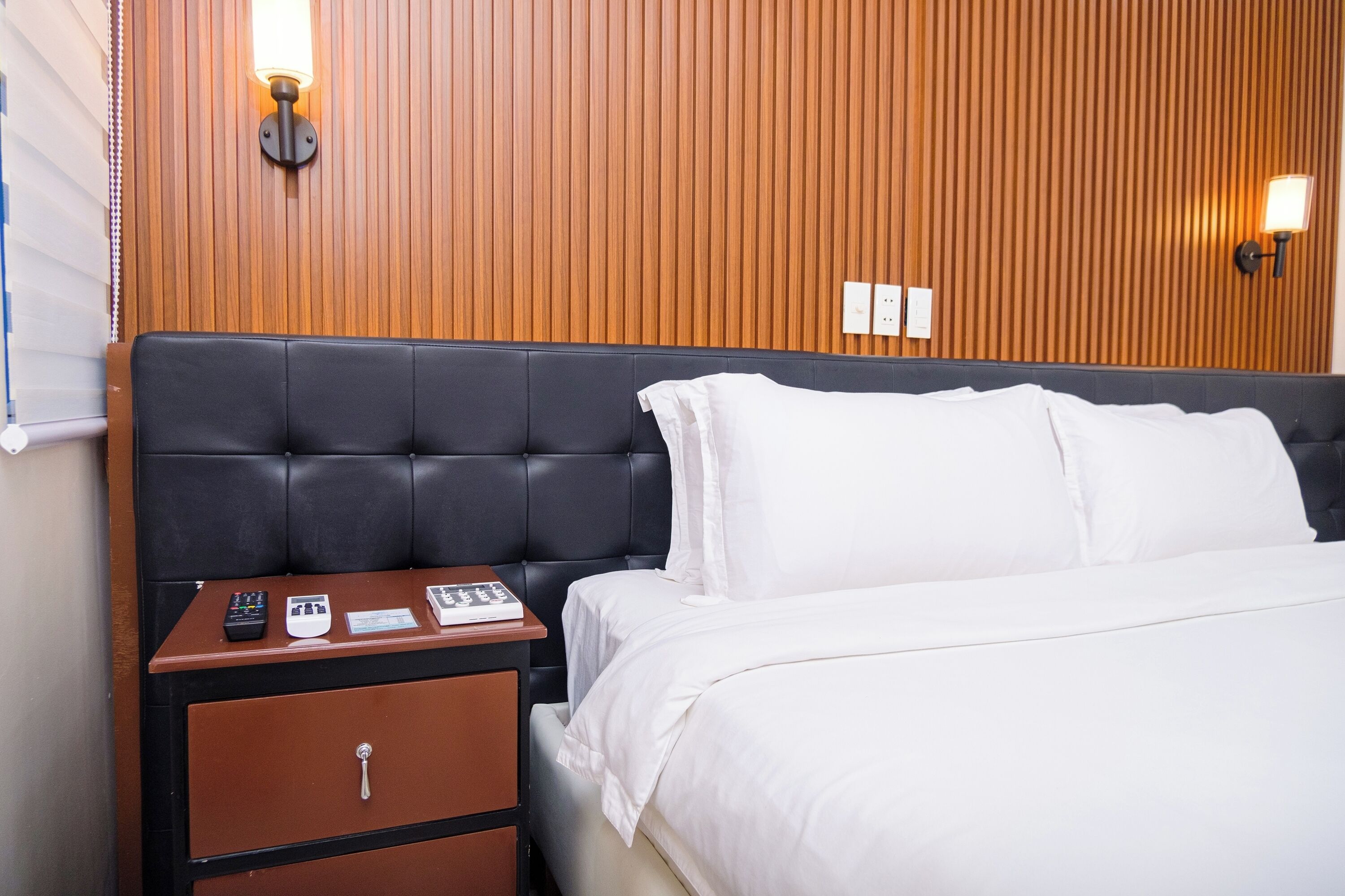 Habitació d'hotel amb un capçal de llit tapissat de color negre, roba de llit blanca, una tauleta de nit de fusta amb un calaix i dues làmpades fixes a la paret.