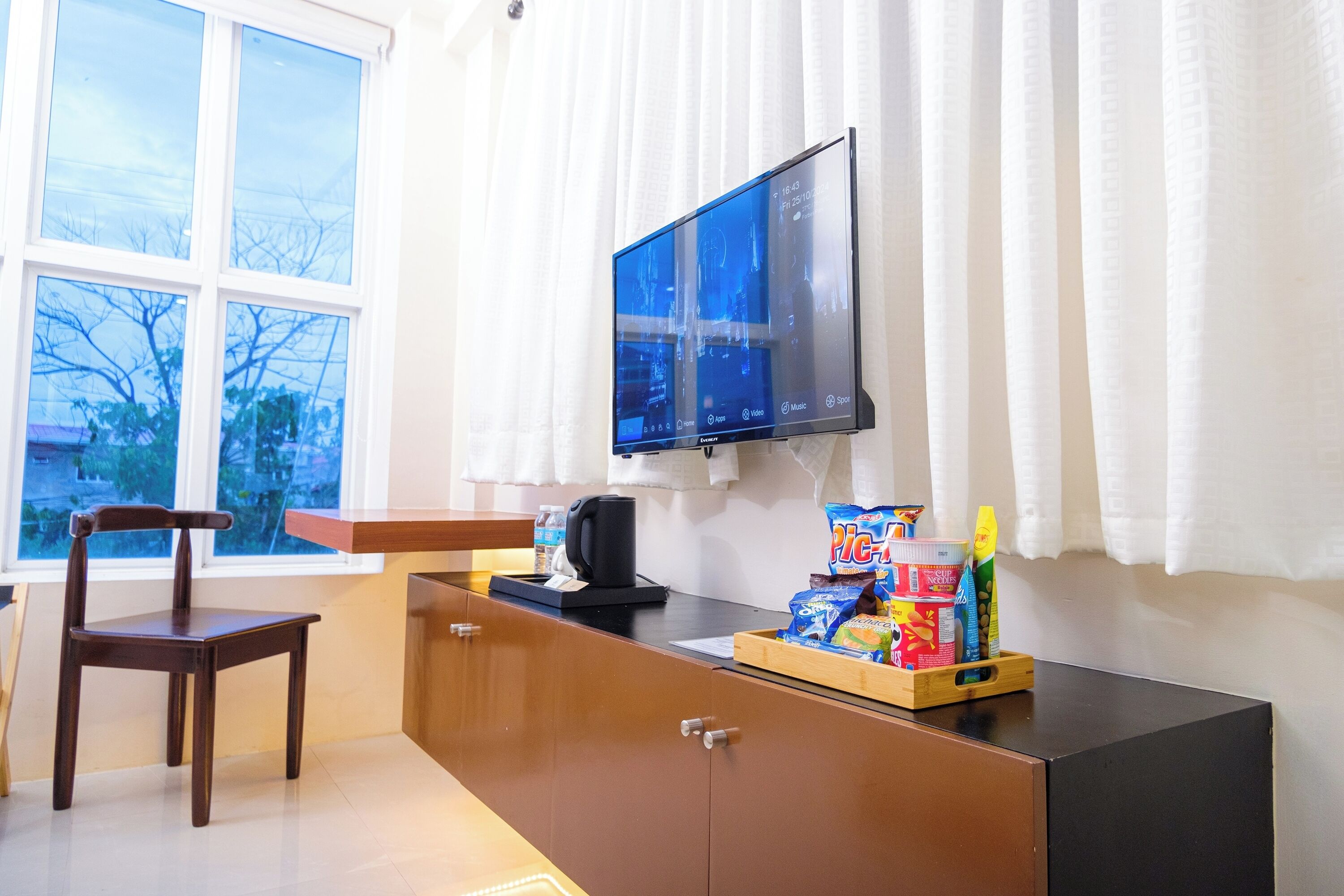 A room with a wooden desk, a chair, a television mounted on the wall, and a window with curtains.