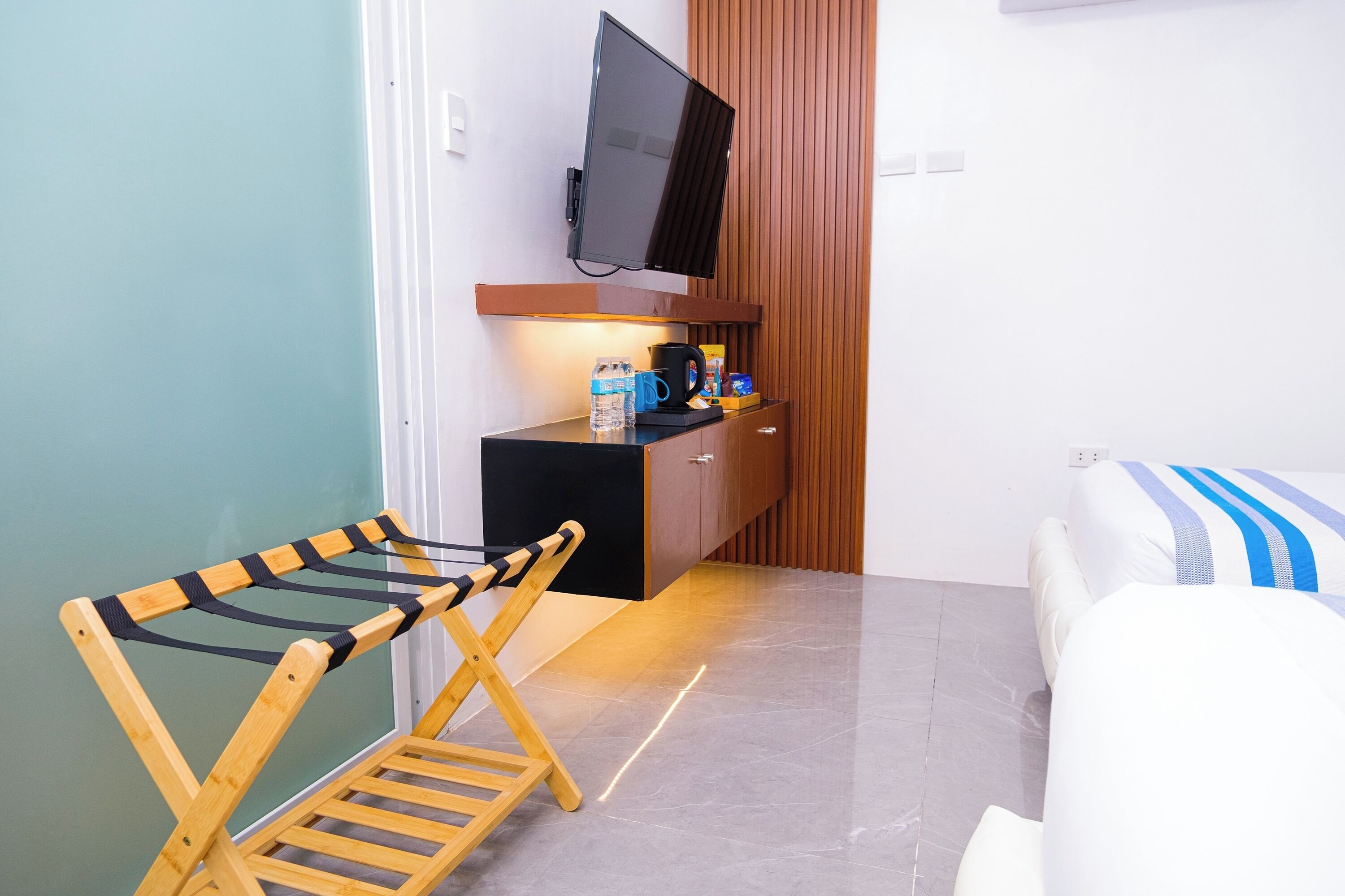 A compact room with a bed, a wooden chair, a small table with a kettle, and a TV mounted on the wall.