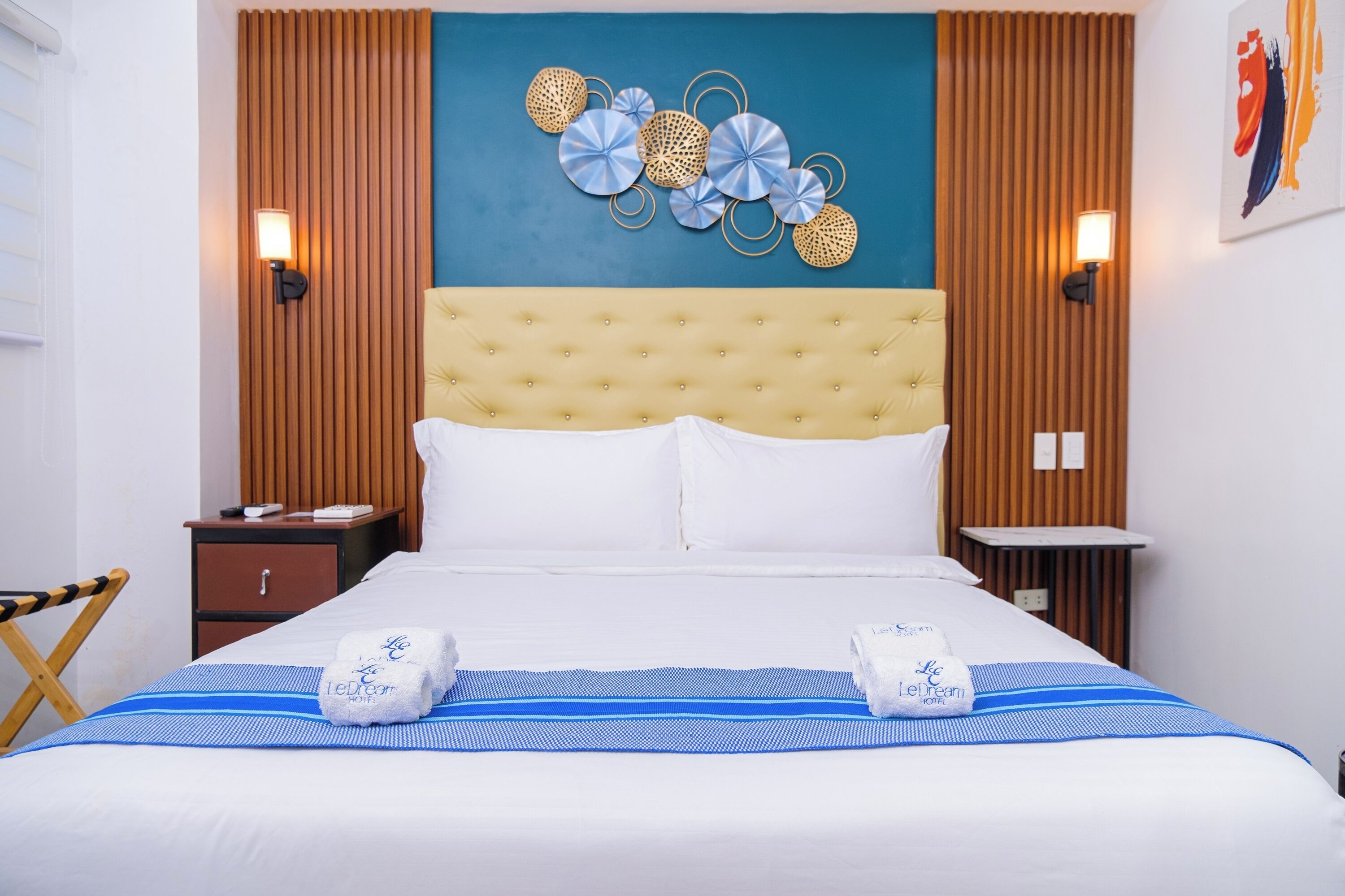 A hotel room with a tufted headboard, a bed with white linens, two towels with a logo, a blue-striped bedspread, and a wall art piece.