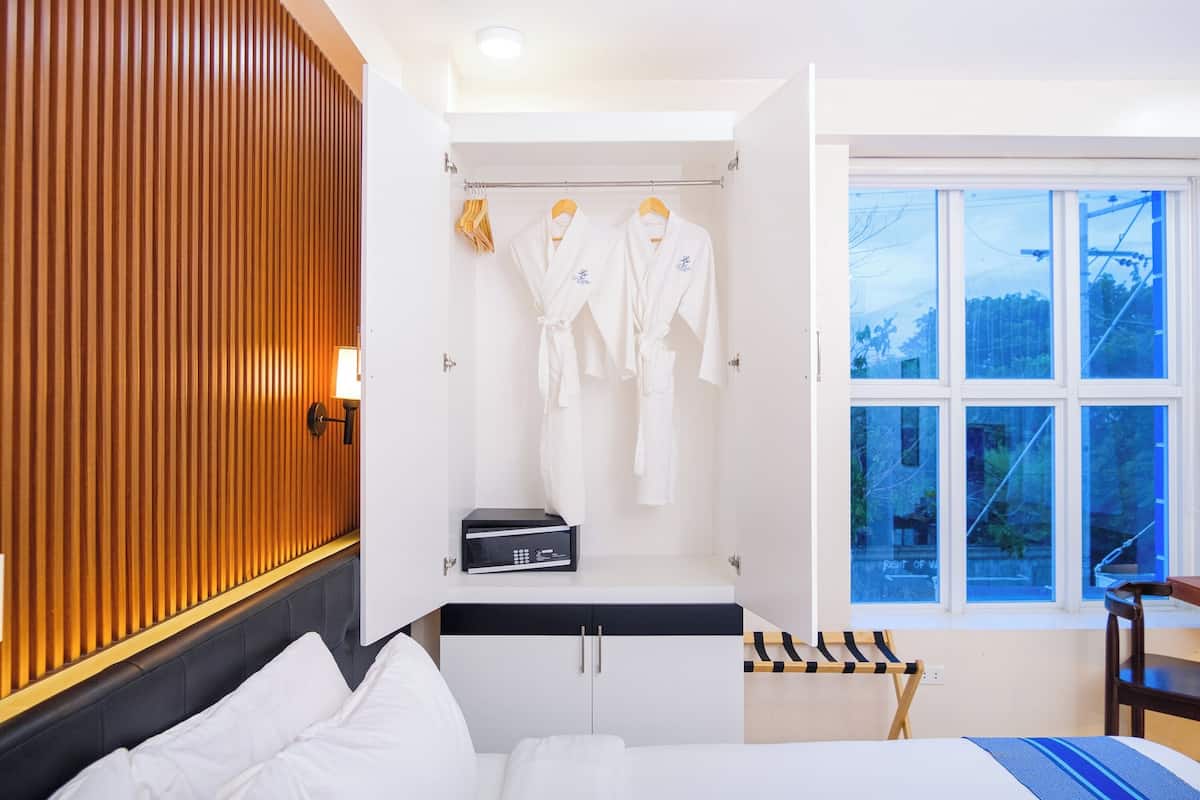 A hotel room with a bed, a wardrobe, a window with a view, and two white robes hanging on a rack.