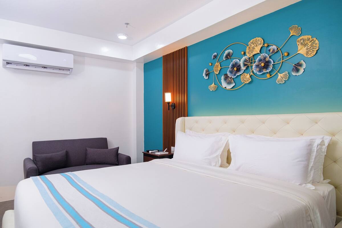 A hotel room with a bed, a chair, a wall-mounted air conditioner, and a decorative wall piece.