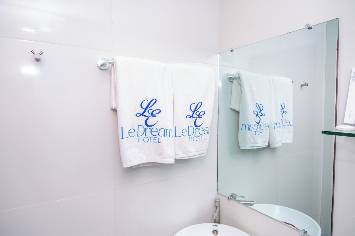 A bathroom with white tiles, a glass shower enclosure, and two white towels hanging on the wall with "LeDream LeDream Hotel" embroidered in blue.