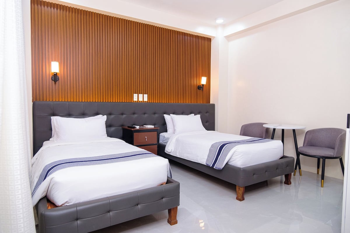 A hotel room with two beds, a small table, a chair, and a wooden panel wall with wall sconces.