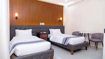 A hotel room with two beds, a small table, a chair, and a wooden panel wall with wall sconces.