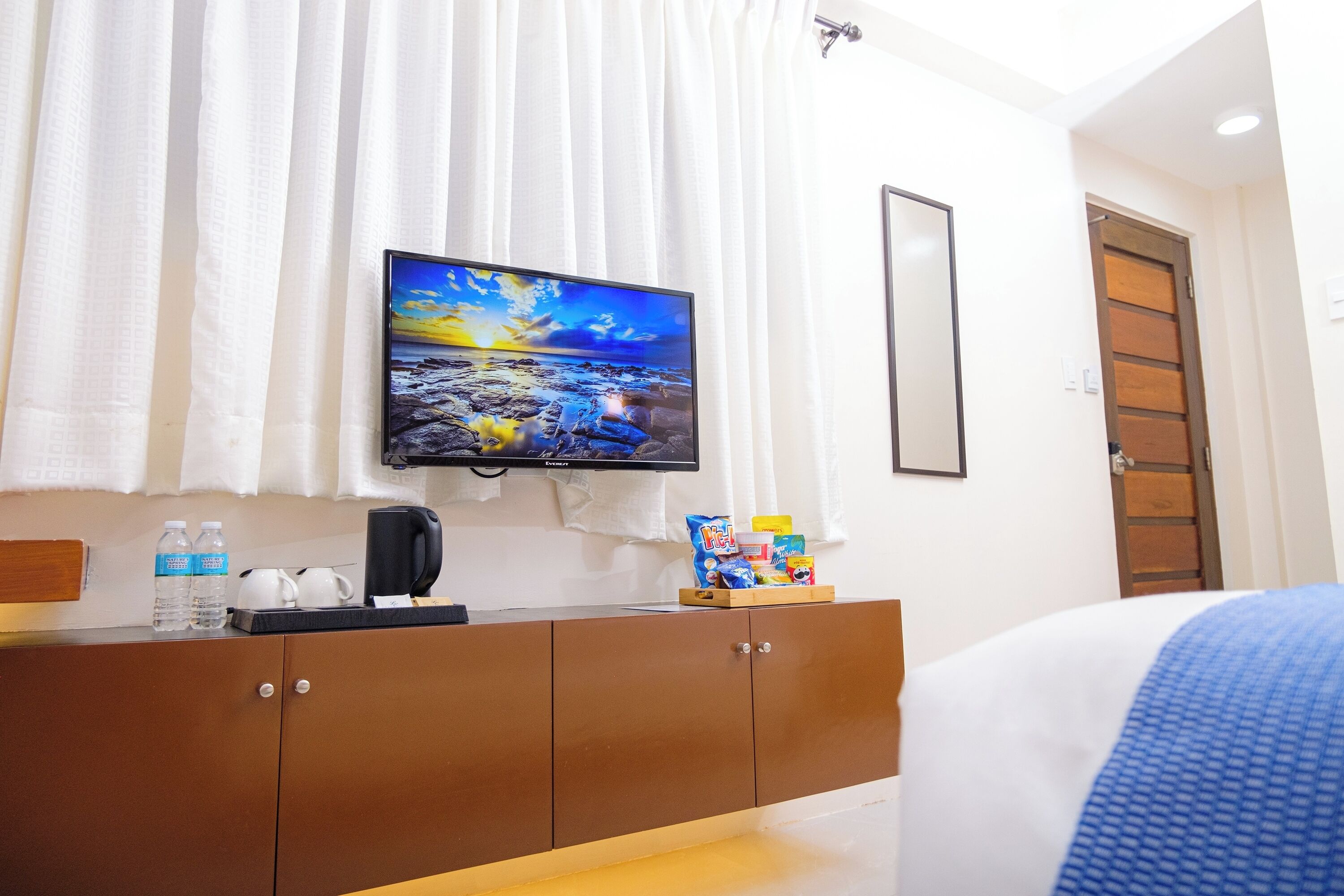 Una habitación de hotel con un televisor montado en la pared, un mueble de madera con artículos de tocador y una cama con una manta azul.