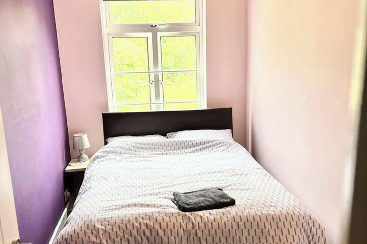A bedroom with a bed, a nightstand, a lamp, and a window with a view of greenery.