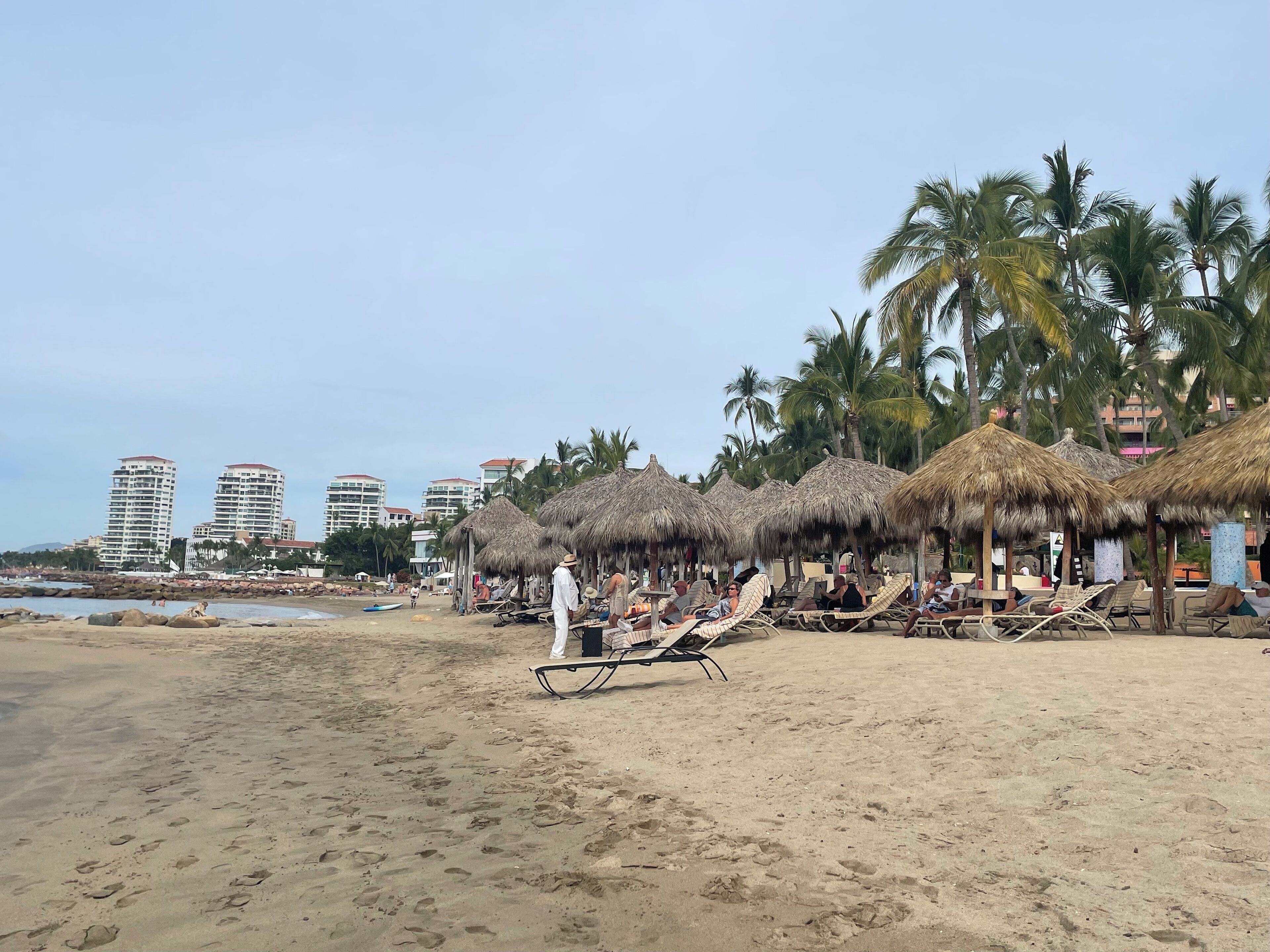 A beach with thatched umbrellas, lounge chairs, and palm trees.