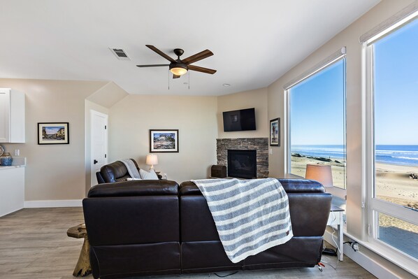 A living room with a leather sofa, a fireplace, a television, and a large window overlooking a beach.