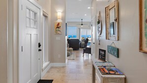 A hallway with a white door, a sign reading "Coastal Bungalow," and a view of a living area with a sofa and a view of the ocean.