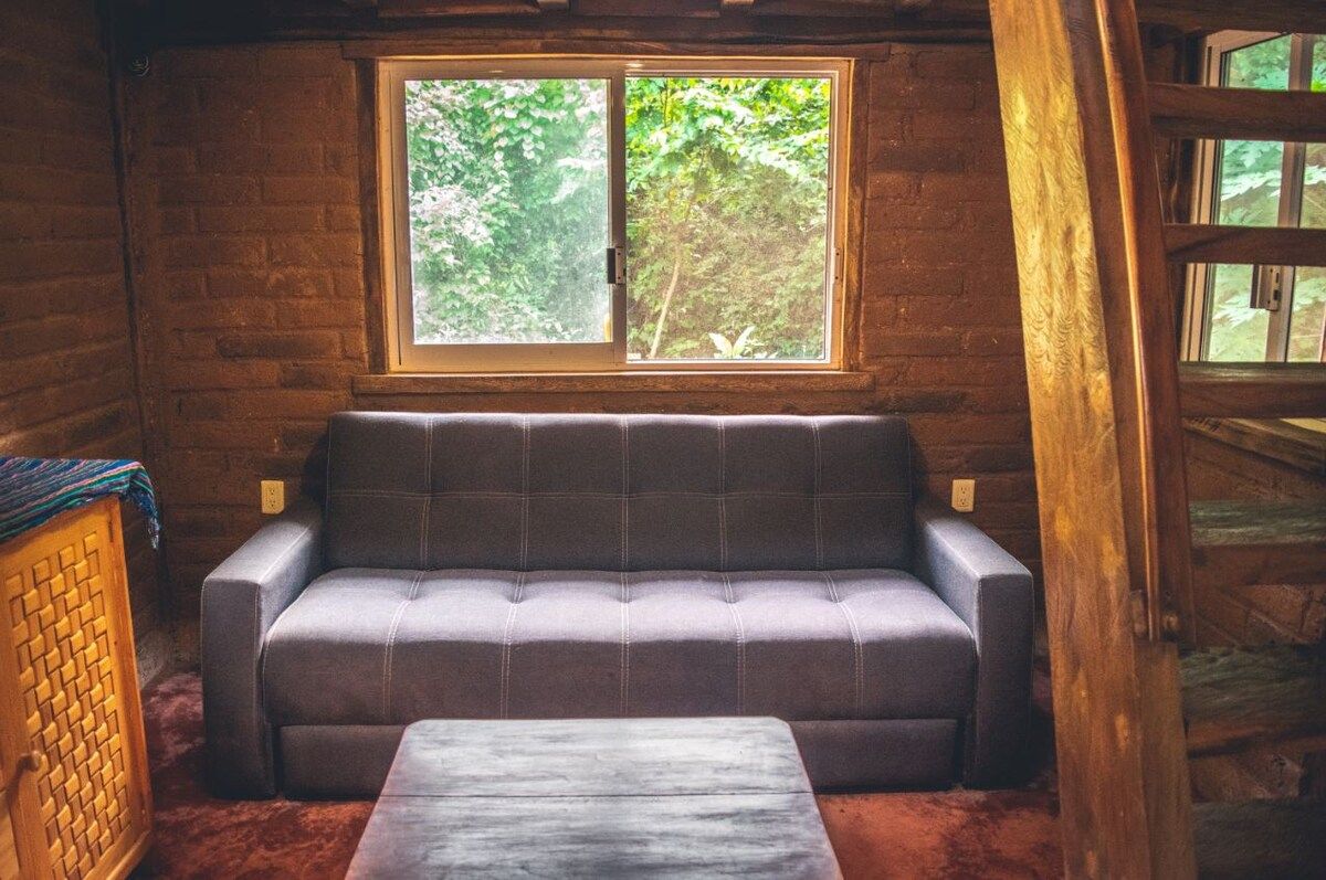 L'intérieur d'une cabane en rondins, avec un canapé et une table, une fenêtre donnant sur un paysage verdoyant et des murs en bois.