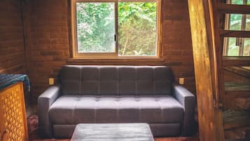 A log cabin interior with a sofa and a table, a window with a view of greenery, and wooden walls.