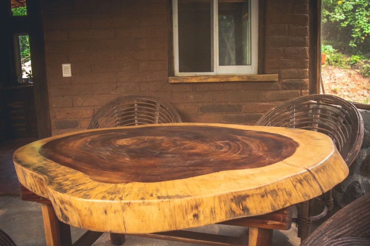 A round wooden table with a central cut-out, surrounded by wicker chairs, set against a brick wall with a window.