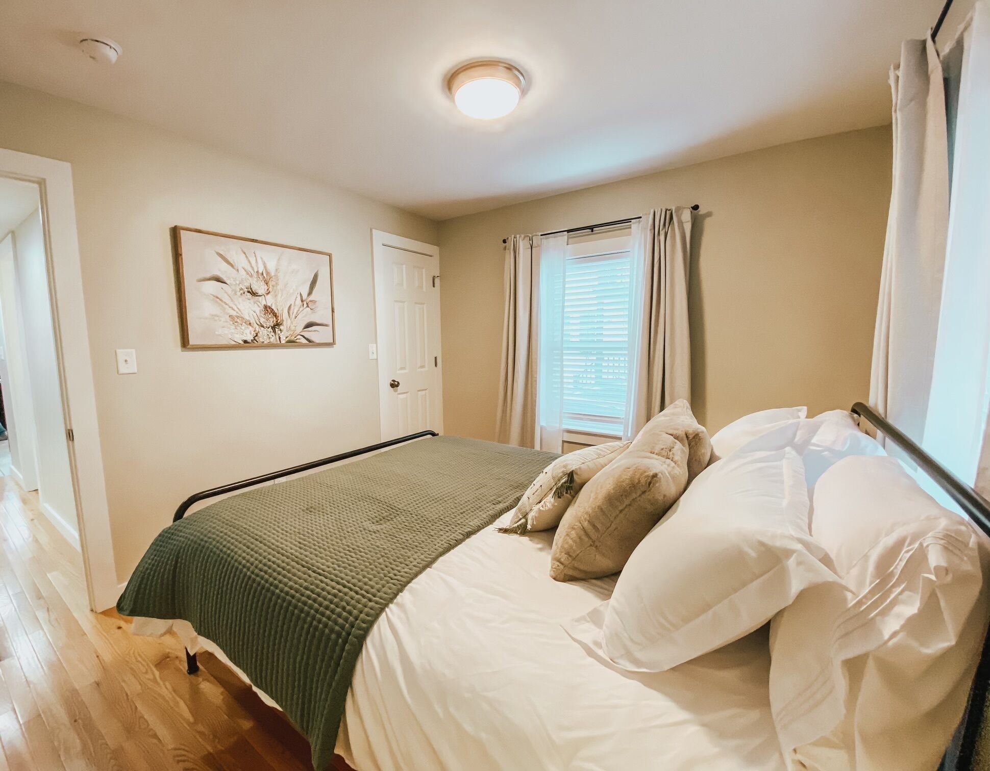 Un dormitorio con cama, manta verde, almohada blanca, almohada color beige, un cuadro en la pared, ventana con cortinas y una puerta.