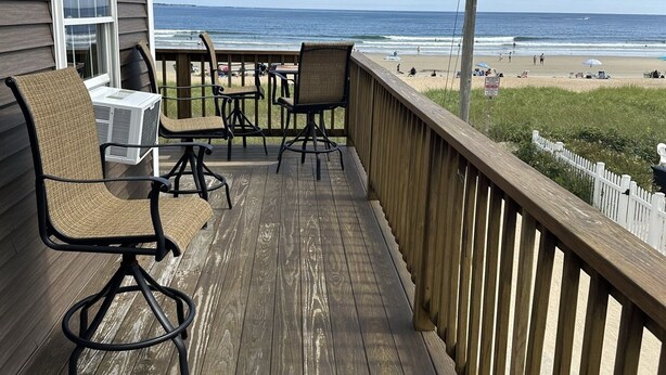Une terrasse en bois, agréée de mobilier d’extérieur, qui donne sur une plage animée et est bordée d’une clôture blanche.