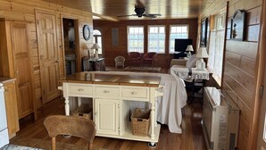 Un intérieur de chalet chaleureux avec des murs en bois, un îlot central doté de tiroirs, des paniers en osier, un ventilateur de plafond et une grande fenêtre.