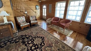 L'intérieur d'un chalet en bois, avec un escalier, deux fauteuils à motifs floraux, un fauteuil en osier et un grand tapis à motifs.