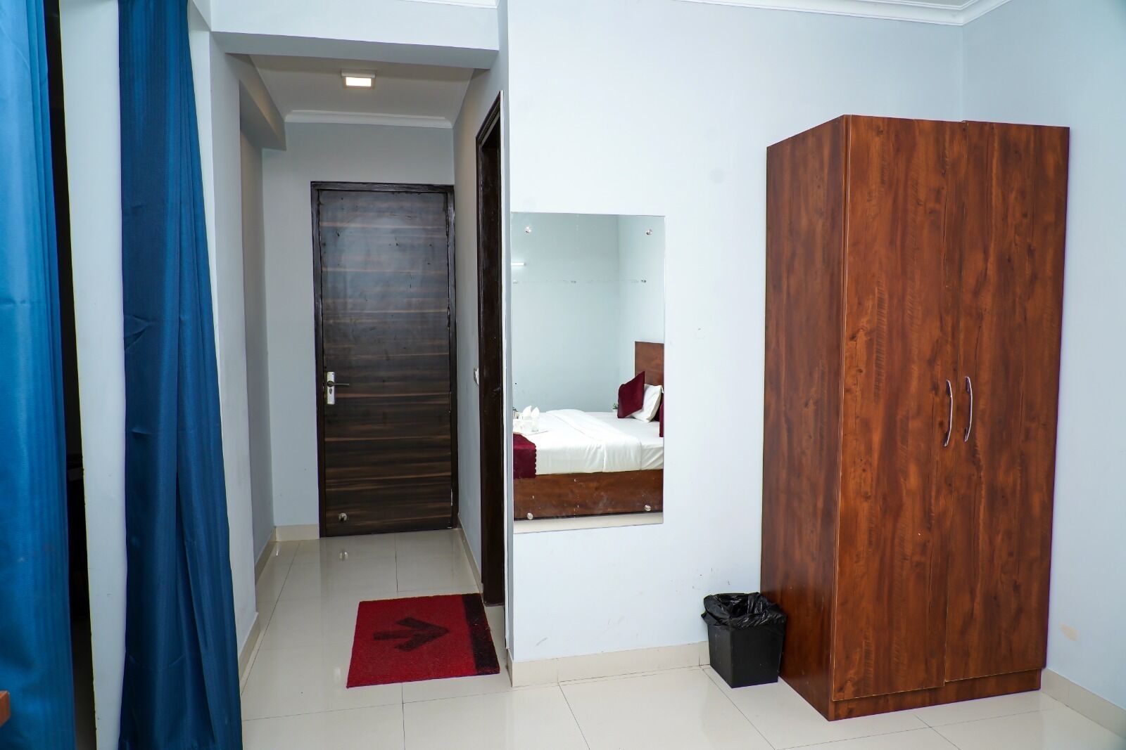 A hotel room with a wooden wardrobe, a bed with white bedding, a red rug, and a small trash bin.