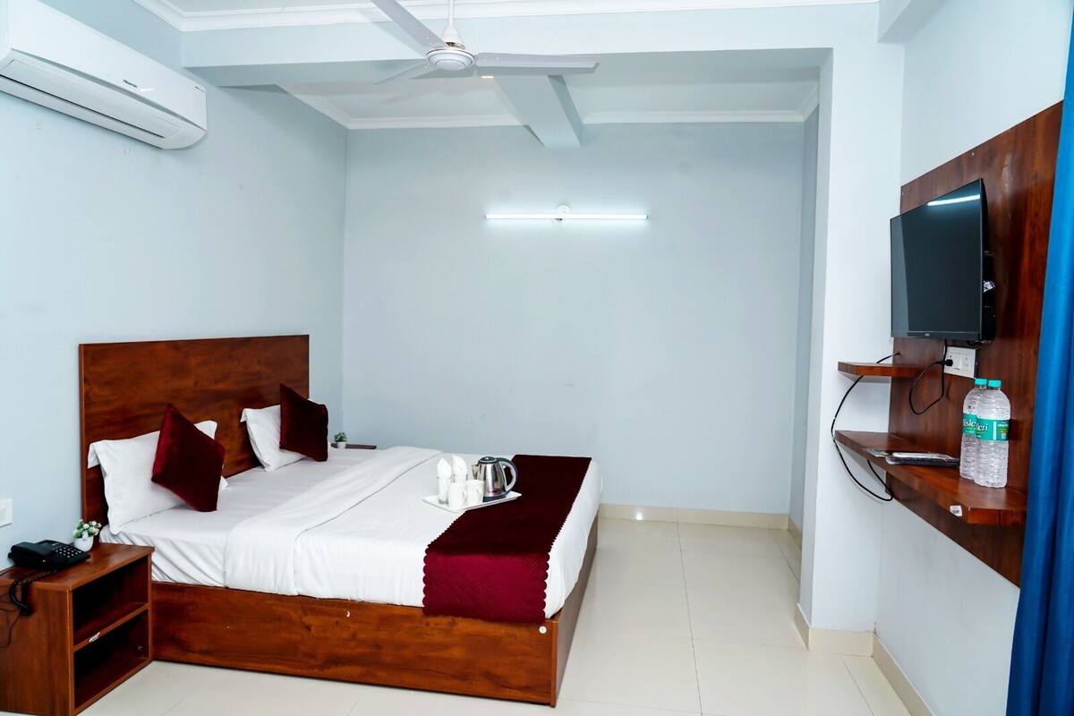 A hotel room with a wooden bed, a bedside table with a phone, a flat-screen TV mounted on a wooden panel, and a water bottle on a shelf.