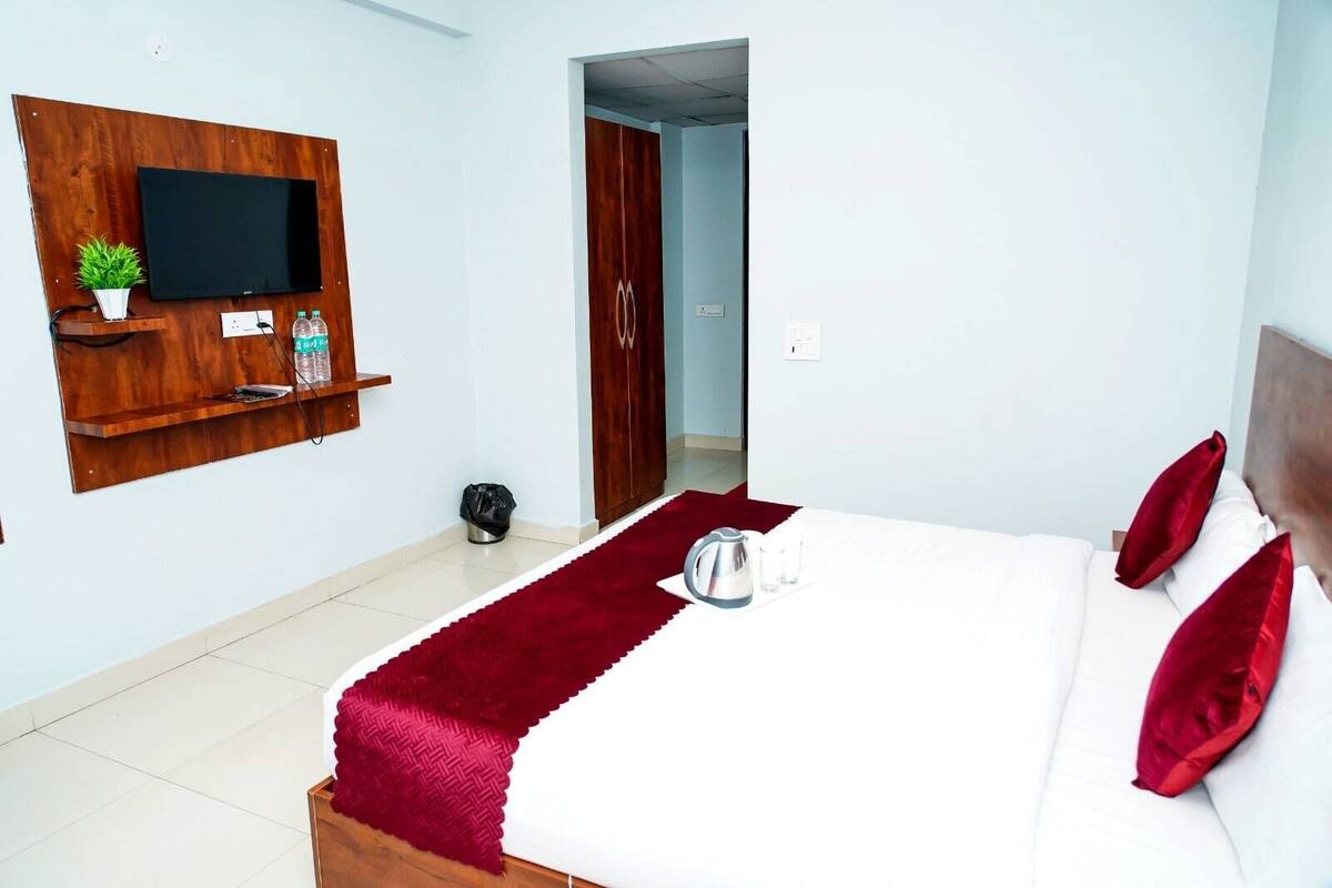 A hotel room with a bed, a TV mounted on a wooden panel, a small plant, a red throw, and a kettle on the bedside table.