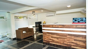A reception area with a wooden reception desk, a brown sofa, a black refrigerator, and a ceiling light fixture. - Shaan Stay (Gurugram)