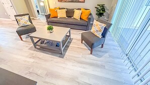 A modern living room with a grey sofa, two armchairs, and a coffee table. There is a painting on the wall and a plant on the right. - Modern Home w/Pool close to Disney Florida theme parks (Kissimmee)