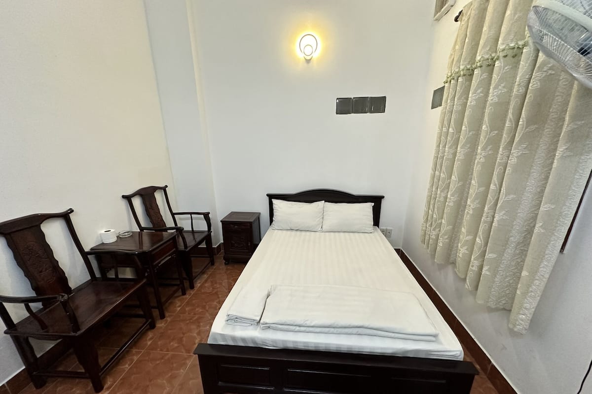 A neatly arranged bedroom with a bed, a small table, two chairs, a wall-mounted light, and a window with curtains.