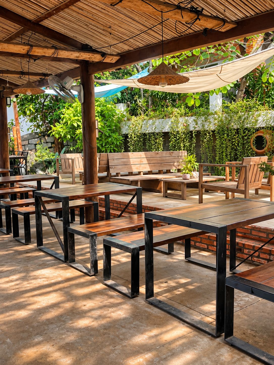 An outdoor patio area with wooden tables and benches, surrounded by greenery and a stone wall.