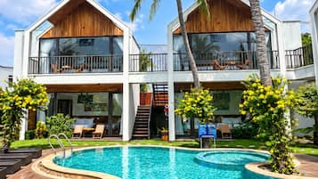 Een resort met twee verdiepingen, een zwembad, palmbomen en een helderblauwe lucht.