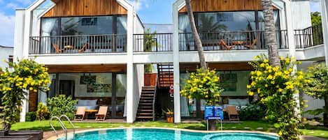 A two-story resort with a swimming pool, palm trees, and a clear blue sky.