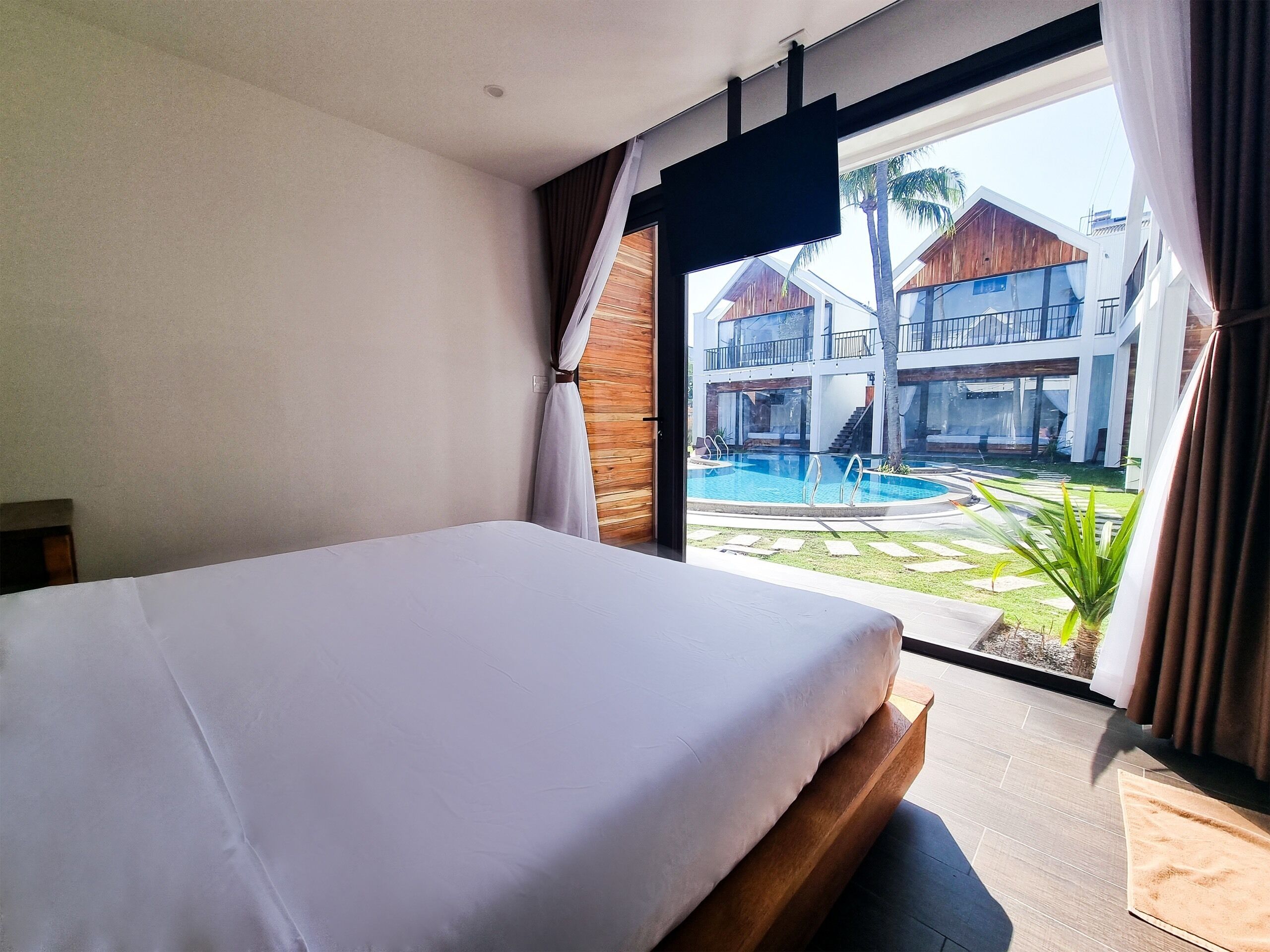 A hotel room with a bed, a window with curtains, and a view of a pool and buildings.