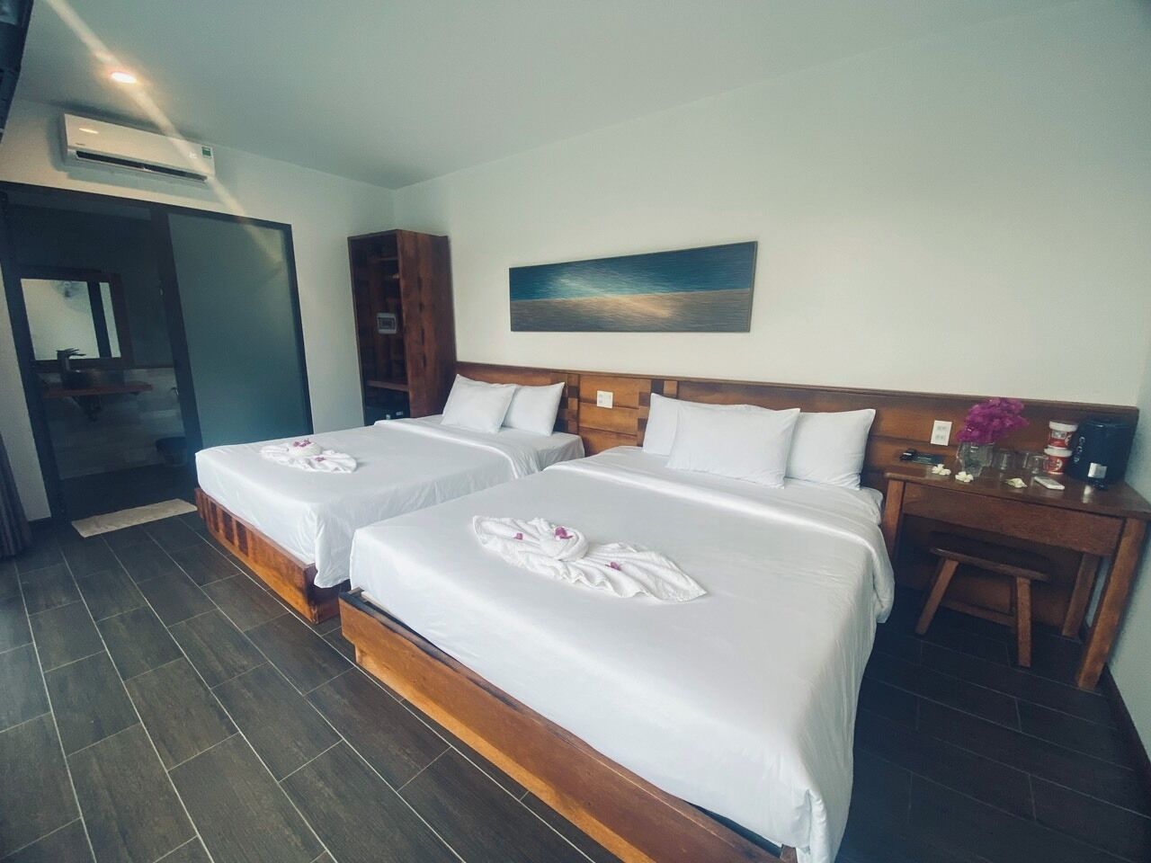 Une chambre d’hôtel avec deux lits, une tête de lit en bois, un tableau au mur et une petite table sur laquelle est posé un vase de fleurs.