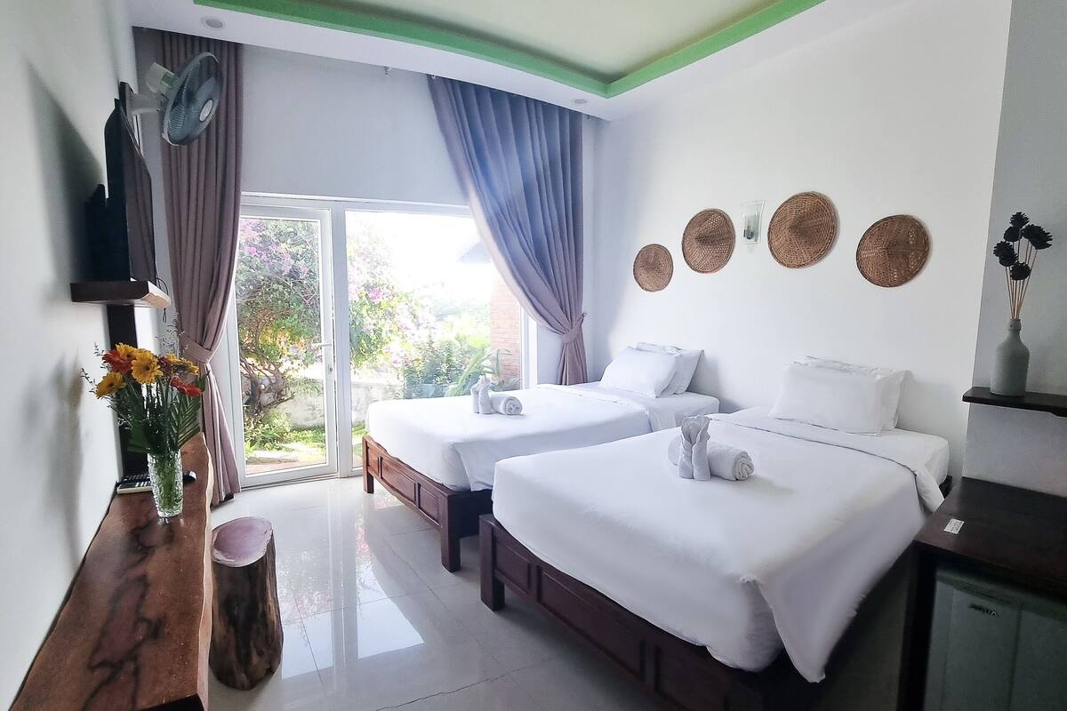 A hotel room with two beds, a wooden bench, a vase with flowers, and a view of greenery outside.