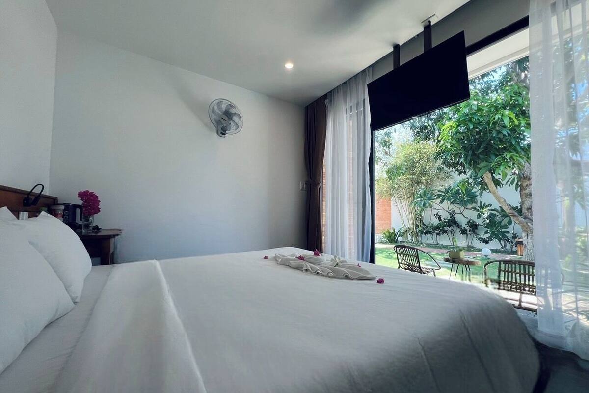 A hotel room with a large bed, a small table with a fan, and a view of the garden through the window.