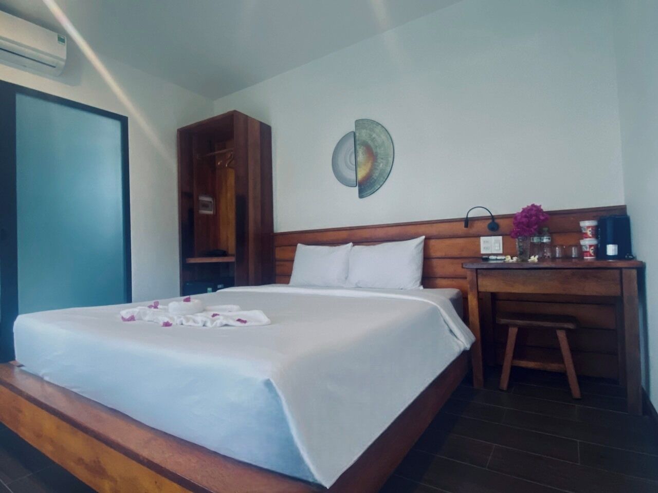 Een netjes opgemaakt bed met een witte deken en een bloemstuk, een houten bureau met een stoel, en een wandplank met diverse spullen.