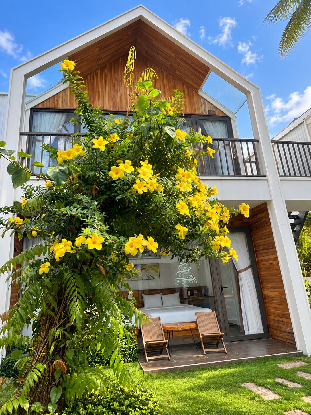 Une maison moderne à deux étages, dotée d’un balcon, d’un jardin parsemé de fleurs jaunes et d’une terrasse avec deux chaises.