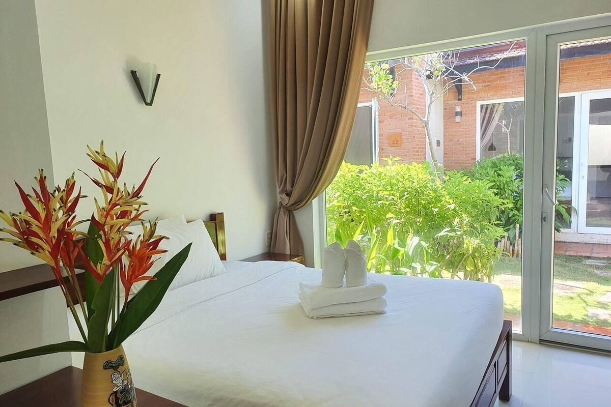 A hotel room with a bed, a vase with flowers, and a view of greenery outside.