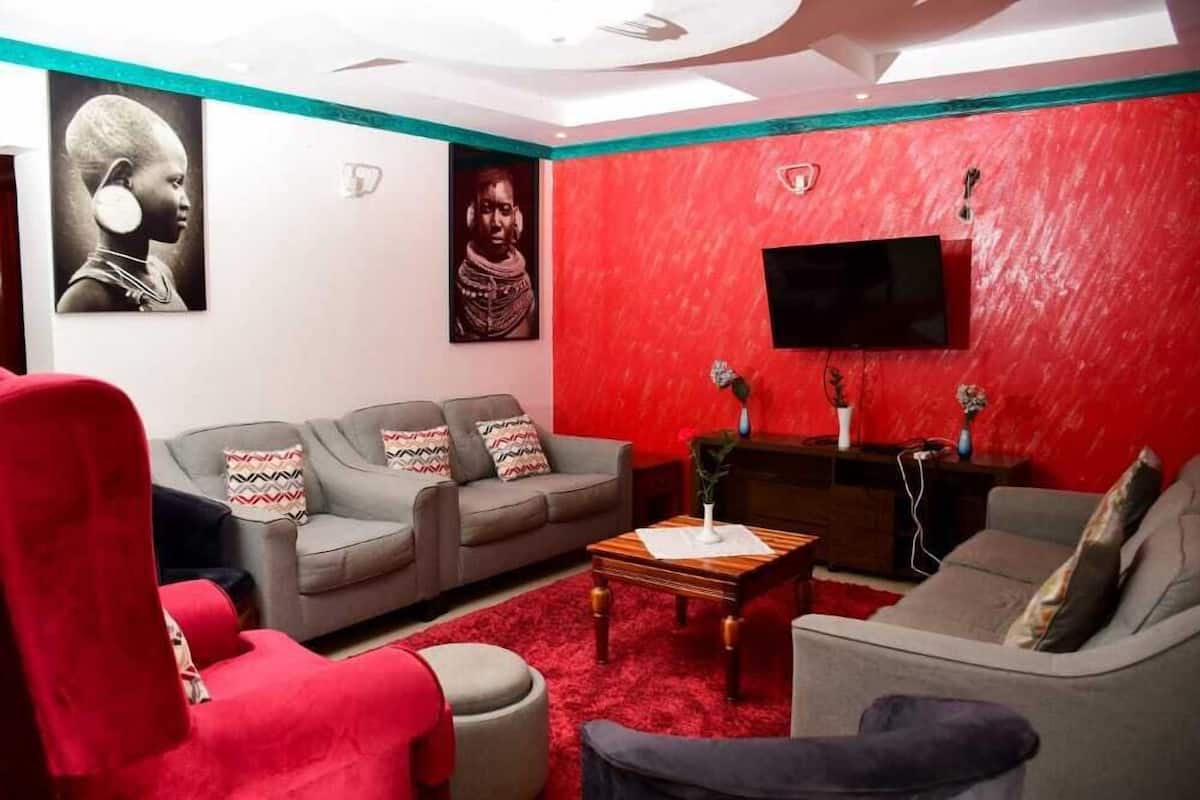 A living room with a red accent wall, grey sofas, a red armchair, and a flat-screen TV mounted on the wall.