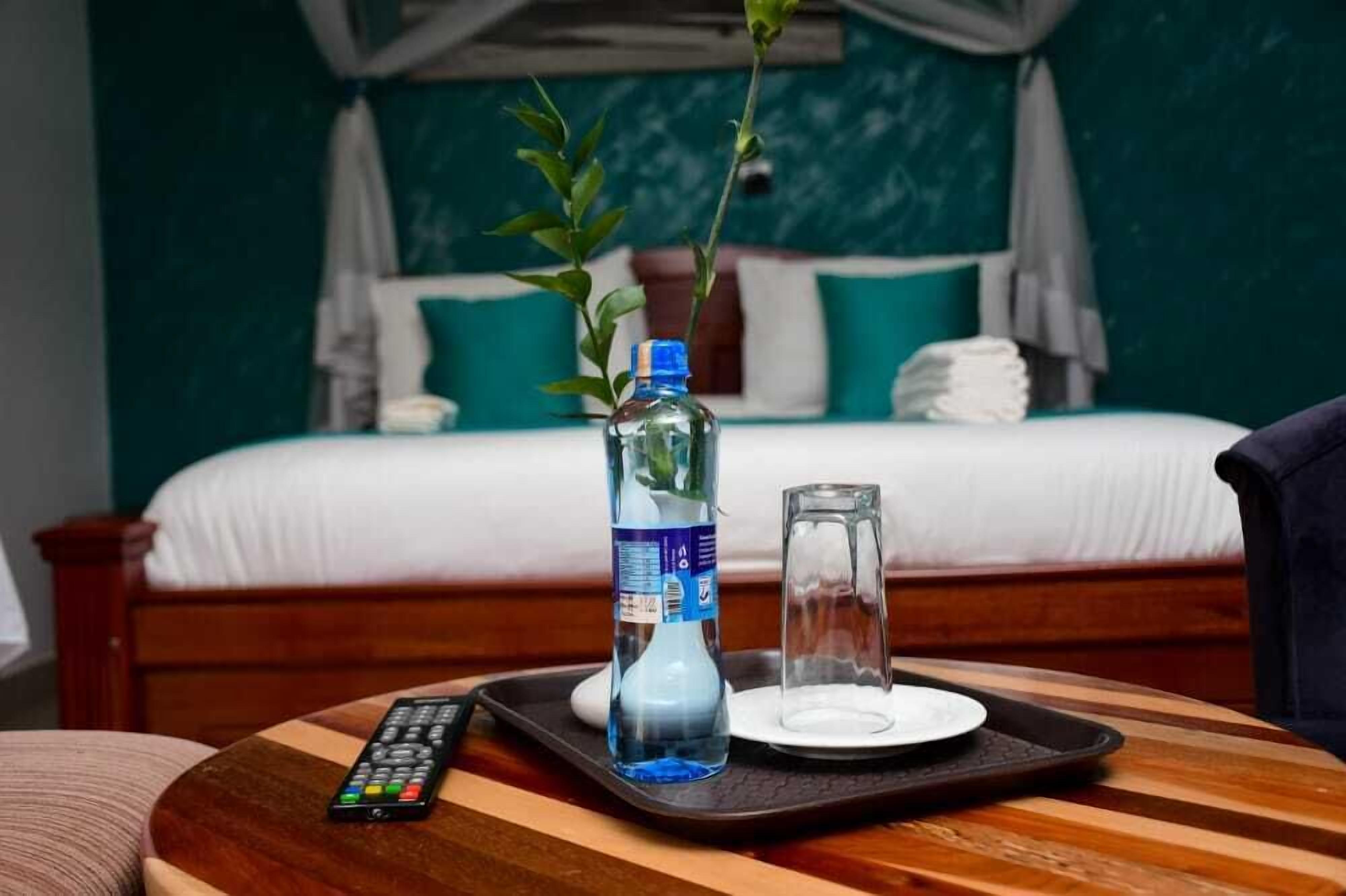 A hotel room with a bed, a bedside table with a remote control, a bottle of water, and a glass.