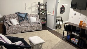 A living room with a floral sofa, a small white ottoman, a black chair, a TV, and a shelf with various items. - Cozy lake house on Sabattus Pond 2 bed 1 Bath  (Sabattus)