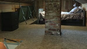 An attic bedroom with a sloped ceiling, a brick pillar, a bed, a desk, and a green folding chair. - Cozy lake house on Sabattus Pond 2 bed 1 Bath  (Sabattus)