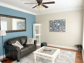 A living room with a black leather sofa, a white coffee table, a ceiling fan, a large mirror, a cabinet, and a floral painting on the wall.