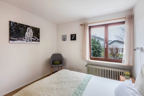 Quarto com cama, janela com cortinas, radiador, cadeira e arte nas paredes.
