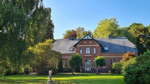 A large brick building with a symmetrical facade, a central entrance, and a well-maintained garden with a stone fountain. - Apartment 'Haloi' with Private Terrace, Private Garden and Wi-Fi (Mittelangeln-Torsballig)