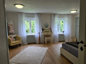 Une chambre à coucher comprenant un lit, une coiffeuse avec un miroir, une chaise, une commode et deux fenêtres avec des rideaux.