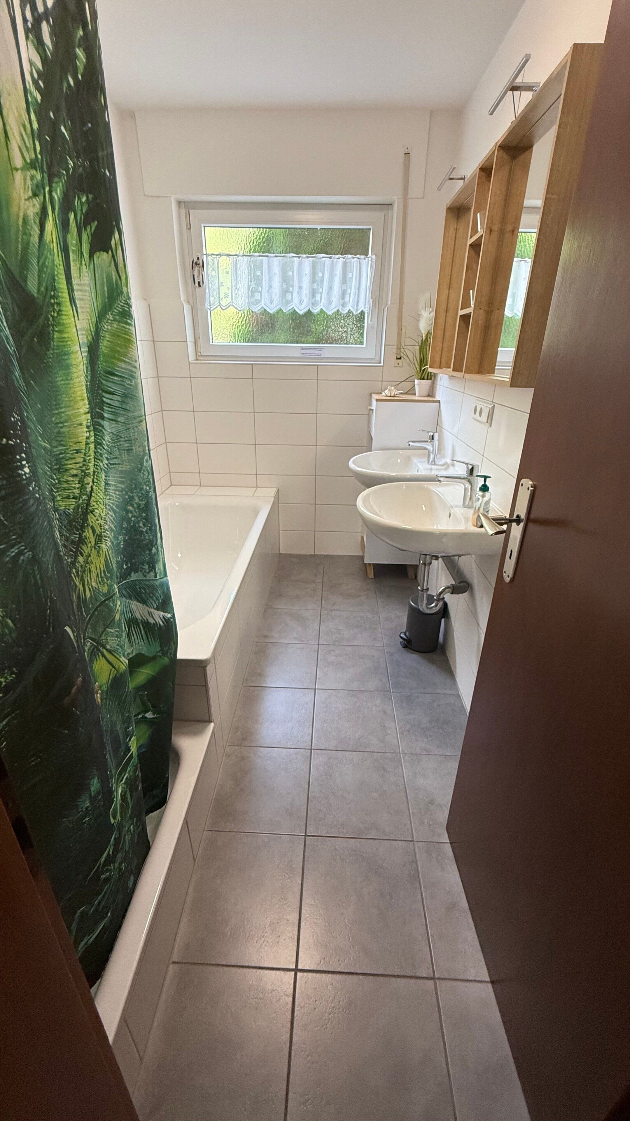 Een badkamer met een badkuip, twee wastafels, een raam met uitzicht en een douchegordijn met een tropisch motief.