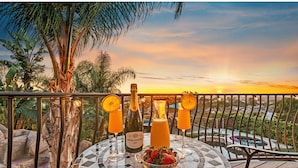 Une vue sur le coucher de soleil depuis un balcon, avec une table dressée pour deux personnes, comprenant une bouteille de vin Piper Sonoma Brut, deux verres de jus d’orange et un bol de fraises.